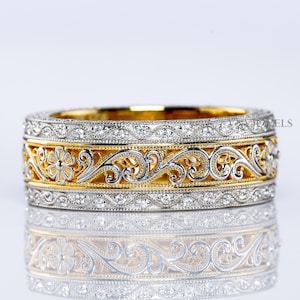 Op de afbeelding: Een tweekleurige ring met ingewikkelde bloemen- en krulontwerpen. De ring heeft een gouden band met een gedetailleerd patroon en een zilveren band met kleine, sprankelende stenen. Het ontwerp van de ring wordt weerspiegeld in het oppervlak.
