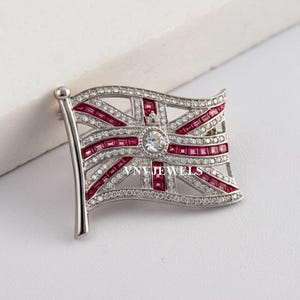 May include: A silver brooch in the shape of a waving Union Jack flag. The flag is decorated with red and clear cubic zirconia stones.