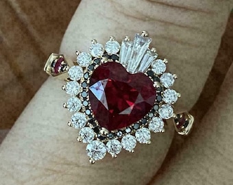 Könnte beinhalten: Ein goldener Ring mit einem herzförmigen, tiefroten Edelstein im Zentrum, umgeben von weißen und schwarzen Edelsteinen. Rote Akzente zieren die Ringschultern. Der Ring ist vor einem neutralen Hintergrund abgebildet.