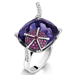 May include: A silver ring featuring a large, square-cut purple gemstone. The ring has a circular design with pink gemstones and a red center. The band is adorned with small, clear gemstones.