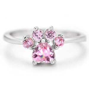 May include: A silver ring featuring a pink paw print design. The paw print is crafted from small pink gemstones with a larger heart-shaped gemstone at its centre.