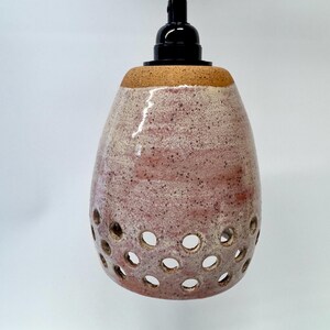 May include: A ceramic pendant light fixture with a speckled pink and cream glaze. The bell-shaped shade features a series of circular cutouts around the bottom edge. It is suspended by a thick jute rope and includes a black plastic socket.