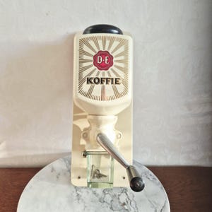 Könnte beinhalten: Eine cremefarbene Wand-Kaffeemühle mit D.E.-Logo und dem Wort KOFFIE in schwarzer Schrift. Das Gerät verfügt über eine metallische Handkurbel, einen schwarzen Deckel und einen kleinen, durchsichtigen Glasbehälter am Boden für das Kaffeemehl.