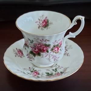 May include: White teacup and saucer set with a floral rose pattern in pink and green. The teacup has a curved handle and gold trim. The saucer has a scalloped edge and gold trim. A classic tea set.
