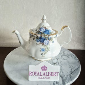 Peut inclure: Une théière en porcelaine blanche ornée d'un motif floral de roses bleues avec des bordures dorées sur le couvercle, l'anse et le bec. Une plaque en céramique blanche affiche le texte violet ROYAL ALBERT ENGLAND avec une icône de couronne.