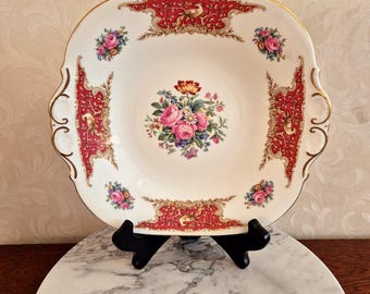 May include: A square white serving plate with a red and gold border and floral design. The plate has two handles on the sides.