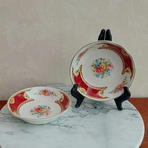 Puede incluir: Dos cuencos de cerámica blanca con un borde rojo y dorado y un patrón floral. Los cuencos están sobre una superficie de mármol blanco.