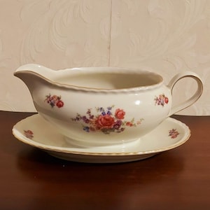 May include: A white porcelain gravy boat with a floral design and a gold trim. The boat is sitting on a matching saucer.