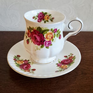 Op de afbeelding: Witte theekop en schotel set met een rozenpatroon in rood, roze en geel. De kop heeft een gebogen oor en staat op een bijpassende schotel. De set is afgewerkt met gouden accenten.