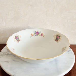 May include: A white porcelain serving bowl with a scalloped edge and gold trim. The bowl is decorated with floral patterns in pink, blue, and yellow. The bowl is on a white marble surface.