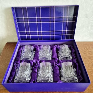 Könnte beinhalten: Ein Set aus sechs Kristallgläsern in einer blauen Geschenkbox. Die Gläser haben ein detailliertes Schliffmuster und liegen auf lila Satin. Die Innenseite des Deckels der Box ist mit einem blauen Karomuster versehen.