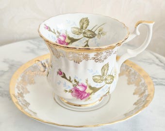 May include: A white porcelain teacup and saucer set featuring a delicate floral pattern of pink roses and green leaves. The rim of both the cup and saucer is decorated with a scalloped gold trim and intricate gold filigree detailing.