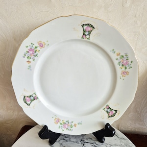 May include: White ceramic plate with a scalloped edge and gold trim. The plate features floral clusters in pink, yellow, and green. Decorative accents are placed around the rim. The plate is displayed on a black stand.