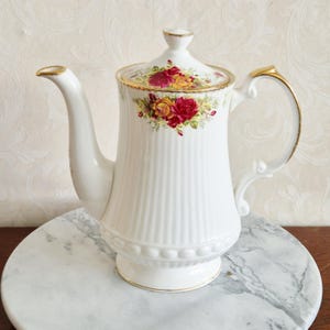 May include: White porcelain coffee pot with a lid, decorated with red and yellow roses. The pot has gold trim on the spout, handle, and base. The body features vertical ribbed detailing. A classic piece for serving coffee.