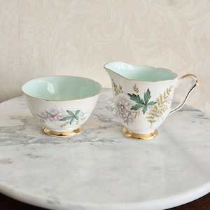 Op de afbeelding: Een vintage porseleinen suikerpot en melkkan set. De witte stukken hebben een bloemenontwerp met roze en groene accenten, gouden randen en een lichtblauwe binnenkant. De set wordt gepresenteerd op een marmeren oppervlak.