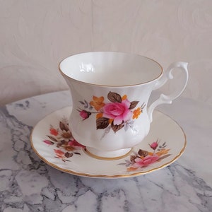 May include: White teacup and saucer set with a floral rose design in pink, orange, and brown. The cup and saucer have gold trim. The teacup has a curved handle.
