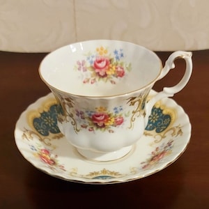 Puede incluir: Una taza de té y un platillo blancos con un patrón floral. La taza y el platillo tienen un borde dorado y un diseño azul y dorado en el platillo.