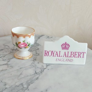 Könnte beinhalten: Ein weißer Porzellan-Eierbecher mit floralem Design und goldenem Rand, neben einem weißen Royal Albert England-Schild mit rosa Kronenlogo und Text.