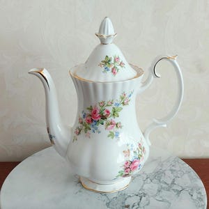 Könnte beinhalten: Eine weiße Porzellan-Kaffeekanne mit einem Blumenmuster aus rosa und blauen Rosen. Die Kanne hat einen goldenen Rand und einen Deckel mit einem Knauf.