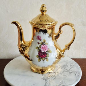 Puede incluir: Una tetera de porcelana dorada con un diseño floral de rosas rosas y rojas. La tetera tiene un pico curvo, un asa de bucle y una tapa con un adorno decorativo. La tetera se exhibe sobre una superficie de mármol.