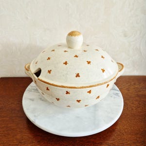 Op de afbeelding: Een crèmekleurige keramische soepterrine met een gespikkelde afwerking en een herhalend patroon van kleine bruine klaverachtige motieven. De terrine heeft een bijpassend bol deksel met een ronde knop, twee zijhandvatten en een inkeping voor een pollepel.