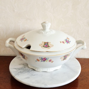 May include: A white porcelain soup tureen with a lid, handles, and a matching base. The tureen is decorated with floral and crest designs in multiple colours, and has gold trim. The tureen is sitting on a round marble base.