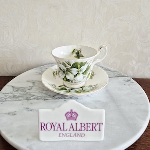 May include: A white teacup and saucer set with a floral design, featuring white flowers and green leaves. The teacup has a gold handle and trim. A small white sign with the text "ROYAL ALBERT ENGLAND" is in front of the teacup.