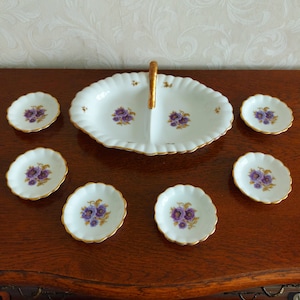 Könnte beinhalten: Eine weiße Porzellanservierschale mit goldfarbenem Rand und einem Griff. Die Schale ist in zwei Abschnitte unterteilt und zeigt ein Blumenmuster mit violetten Blüten. Die Schale ist von sechs kleineren weißen Porzellanschalen mit goldfarbenem Rand und dem gleichen Blumenmuster umgeben.