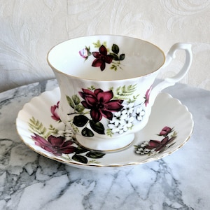 May include: A white teacup and saucer with a floral pattern featuring burgundy and white flowers. The cup and saucer have a gold trim.