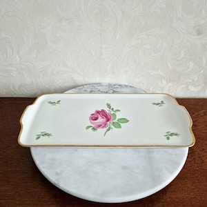 Puede incluir: Una bandeja rectangular de cerámica blanca con ribete dorado y un diseño floral de rosa rosa. La bandeja tiene un borde festoneado y descansa sobre una superficie de mármol.