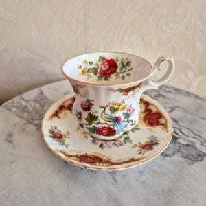 Op de afbeelding: Een wit theekopje en schotel met een bloemen- en vogelontwerp. Het kopje heeft een gegolfde rand en een handvat. De schotel heeft een rode en gouden rand.