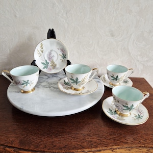 Op de afbeelding: Een vintage theeservies bestaande uit vier kopjes en schotels, plus een extra schotel. De witte porseleinen stukken zijn versierd met een bloemmotief in groen, roze en goud. De kopjes hebben gouden handvatten en bodems. De schotels hebben een diameter van 15 cm.