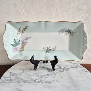 Pode incluir: Bandeja retangular com um design floral azul claro e branco. A bandeja tem uma borda dourada e uma borda ondulada. O design floral inclui folhas verdes e flores rosa e brancas. A bandeja é exibida em um suporte preto.