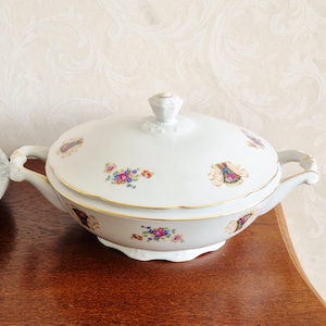 May include: White porcelain serving dish with lid, featuring floral and gold trim details. The dish has two handles and a decorative knob on the lid. The floral pattern includes pink, blue, and yellow flowers.