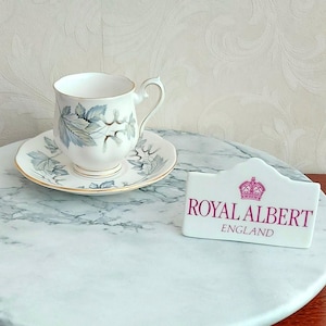 Könnte beinhalten: Eine weiße Teetasse und Untertasse mit blau-grauem Blattmuster und Goldrand. Das Set wird mit einem weißen Royal Albert England-Schild auf einer Marmoroberfläche präsentiert.