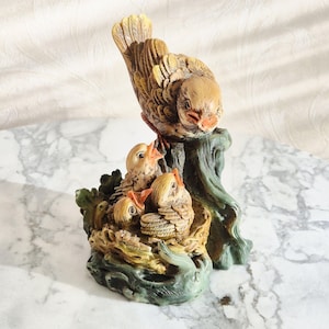 Op de afbeelding: Gedetailleerd beeldje van een vogel die zijn jongen voedt. De volwassen vogel zit op een groene en bruine boomstam, met drie jonge vogels in een nest eronder. Het beeldje is geschilderd in tinten van bruin, bruin en oranje.