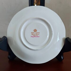 May include: White saucer with a gold rim and a gold inscription that reads "Royal Albert Bone China England Lady Hamilton".