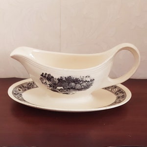 May include: A white ceramic gravy boat with a black and white landscape design. The boat has a matching saucer with a black and white floral border.