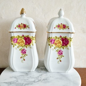 May include: Two white ceramic jars with lids, decorated with a floral rose pattern in yellow, pink, and red. The lids have gold accents, and the jars are displayed on a marble surface. A decorative kitchenware item.