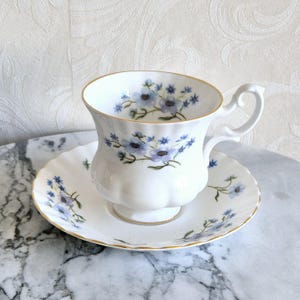 May include: White teacup and saucer set with a floral pattern in shades of blue and purple. The cup and saucer have gold trim. The teacup has a decorative handle and sits on a small pedestal base.