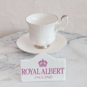 May include: White teacup and saucer with a silver trim. A white sign with a pink crown and the text "Royal Albert England" is in the foreground.