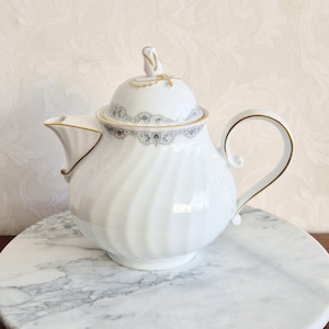 May include: White porcelain teapot with a swirling pattern, lid, and curved handle. The lid features a floral finial and a grey and gold lace design. Gold trim accents the spout and handle, creating an elegant aesthetic. A classic tea set item.