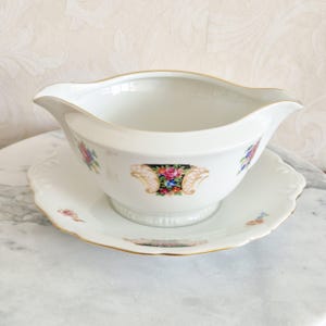May include: A white porcelain gravy boat and saucer set. The gravy boat has a gold rim and a floral design with a black background. The saucer has a scalloped edge and a gold rim. The set is on a marble surface.