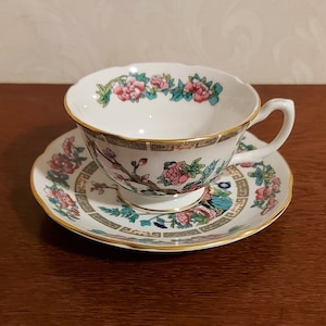 Könnte beinhalten: Eine weiße Teetasse und Untertasse mit einem floralen Muster in Rosa-, Grün- und Blautönen. Das Muster zeigt einen stilisierten Zweig mit Blüten und Blättern sowie einen Vogel mit langem Schwanz. Tasse und Untertasse haben einen goldenen Rand.
