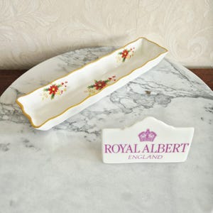Può includere: Un piatto da portata lungo e rettangolare in porcellana bianca con bordi smerlati, finiture dorate e un motivo festivo con stelle di Natale. Accanto si trova una targa in ceramica bianca con un logo a corona viola e il testo ROYAL ALBERT ENGLAND.