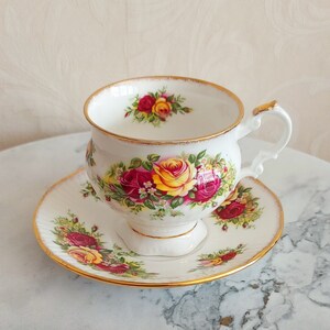 May include: A white teacup and saucer set with a floral pattern featuring red, yellow, and pink roses. The set has a gold trim around the edges.