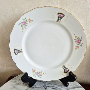 May include: A white porcelain plate with a scalloped edge and gold trim. The plate features floral bouquets in pink, yellow, and blue, along with black and gold decorative accents. The plate is displayed on a black stand.