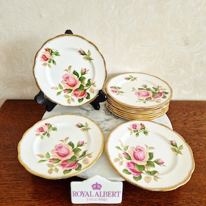 Pode incluir: Um conjunto de pratos de porcelana branca com um design floral de rosas cor-de-rosa e bordas douradas. Os pratos estão dispostos sobre uma superfície de mármore, com um sinal Royal Albert England.