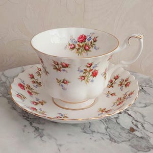 May include: A white teacup and saucer set with a floral pattern of pink roses and blue flowers. The set has a gold trim around the edges.