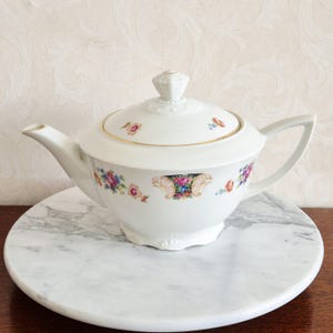 May include: White porcelain teapot with a lid, spout, and handle. The teapot is decorated with floral patterns in pink, blue, and yellow. It sits on a round, marble-patterned surface. The teapot has a gold trim.